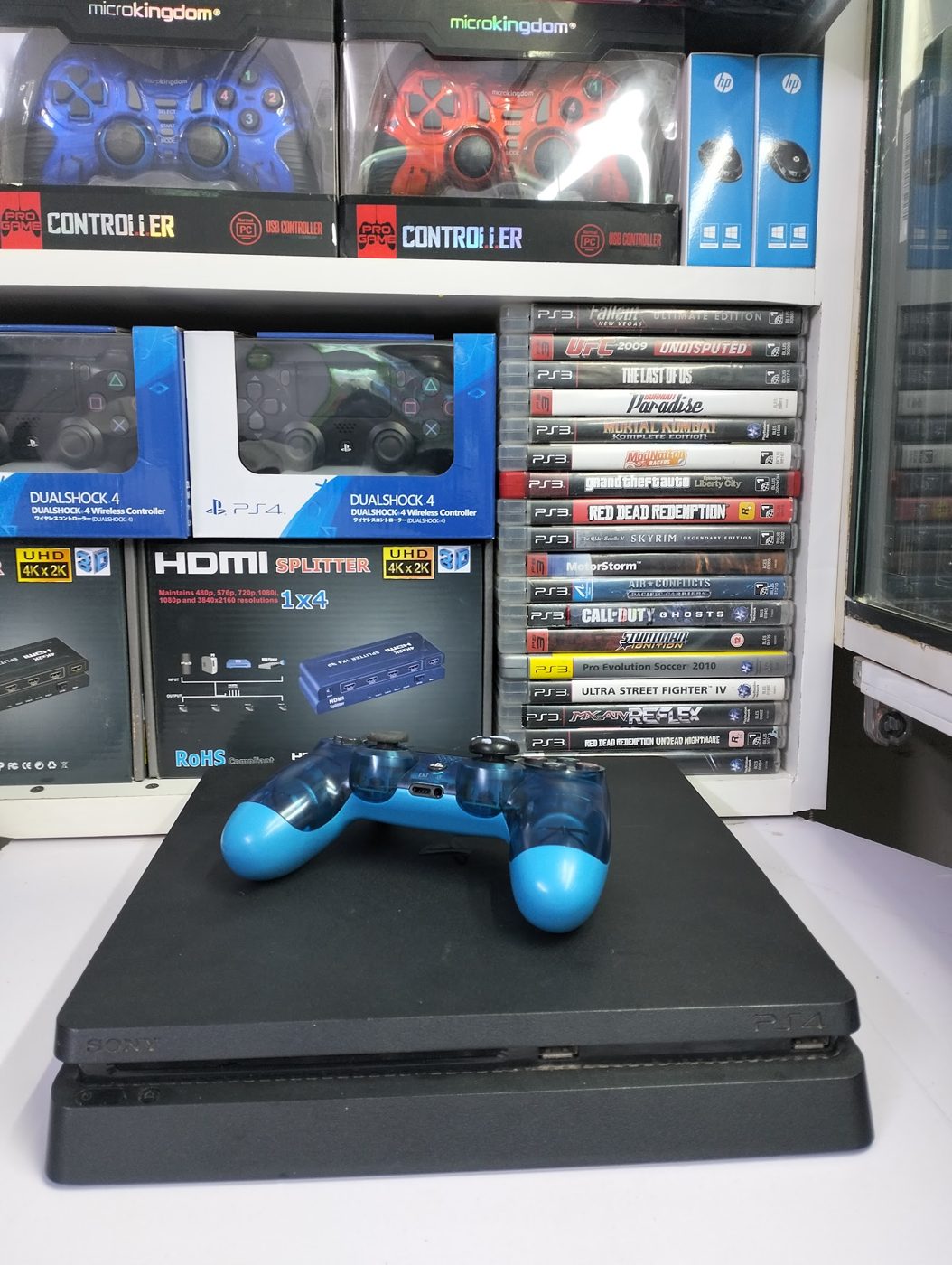 A PS4 Slim with a blue controller, HDMI splitters and pads on the shelf